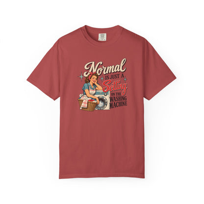 A short-sleeve crewneck t-shirt in a dusty rose or muted brick red color, featuring a vintage-style pin-up graphic of a woman washing clothes with the text 'Normal is just a Setting for the Washing Machine' printed on the front.