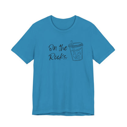 A bright blue short-sleeve crewneck t-shirt with a dark graphic on the chest that reads 'On the Rocks' next to a simple outline drawing of a iced coffee cup.