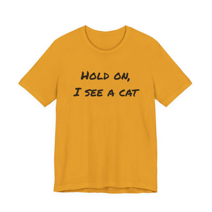 A mustard yellow short-sleeve t-shirt with the text 'HOLD ON, I SEE A CAT' printed in black across the chest, displayed on a solid white background.