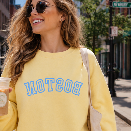 Woman walking in Boston wearing  a Yellow Crew Neck Sweatshirt. Sweatshirt says BOSTON in blue upside down.