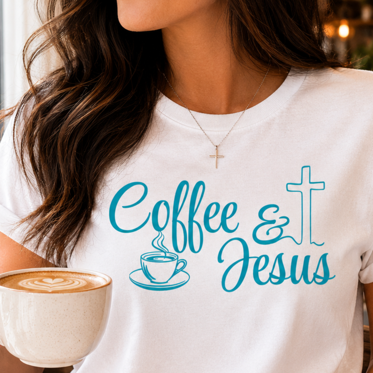 A woman wearing a white crewneck t-shirt with a teal graphic that reads 'Coffee & Jesus' alongside a coffee cup and cross illustration. She is holding a cup of coffee in a warm indoor setting.