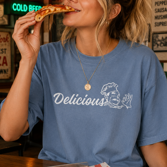 A woman wearing a blue graphic t-shirt with the word 'Delicious' and a chef illustration printed in white on the front, shown as she takes a bite of a slice of pizza in a restaurant setting.