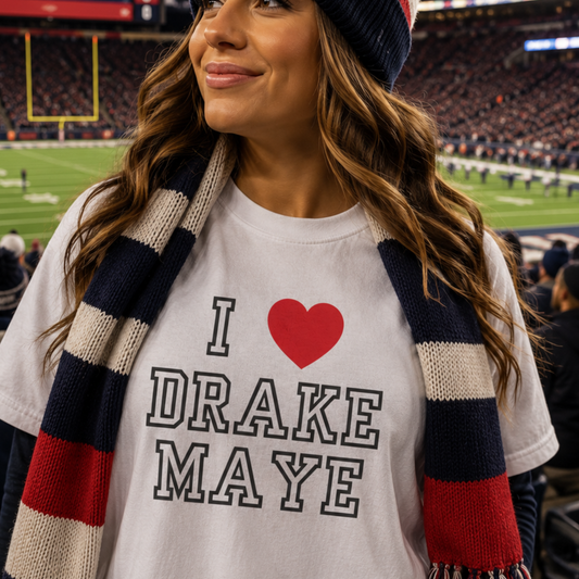 A woman at a football stadium wearing a white graphic T-shirt that reads 'I [red heart] DRAKE MAYE' with a navy, cream, and red striped knit scarf.
