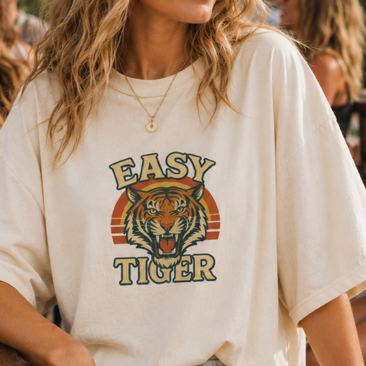 A woman wearing an oversized cream-colored t-shirt with a retro-style graphic of a tiger's face and the words 'EASY TIGER' in yellow and green, set against a warm, blurred background.