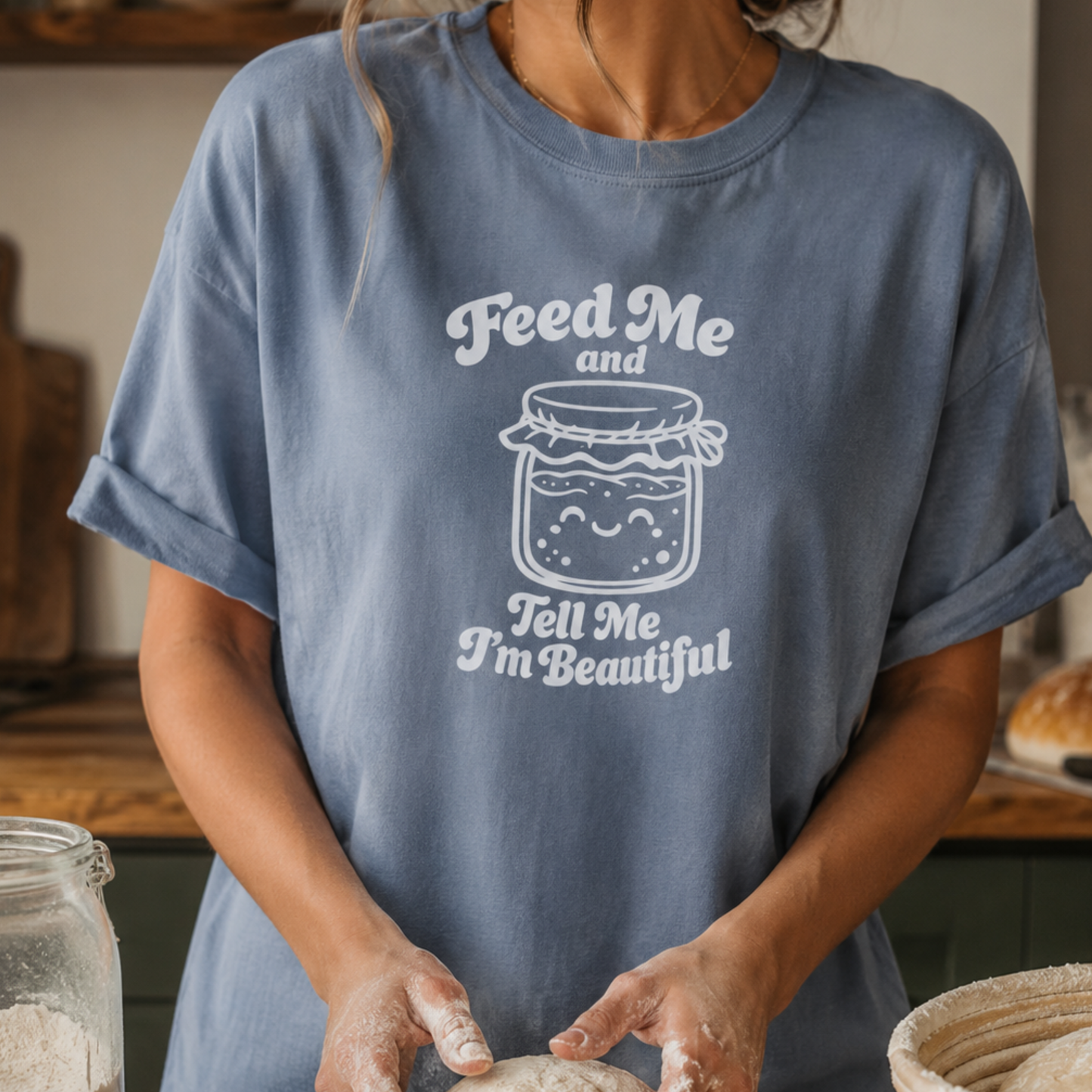A person wearing a dusty blue oversized T-shirt with a white graphic of a smiling jar and the text 'Feed Me and Tell Me I'm Beautiful', standing in a kitchen-like setting with flour visible on their hands.