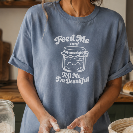 A person wearing a dusty blue oversized T-shirt with a white graphic of a smiling jar and the text 'Feed Me and Tell Me I'm Beautiful', standing in a kitchen-like setting with flour visible on their hands.