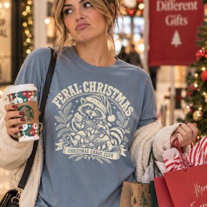 A woman in a festive holiday setting wearing a dusty blue graphic t-shirt that reads 'Feral Christmas Christmas Chaos Club' with a raccoon illustration. She is holding a Starbucks coffee cup and shopping bags, with a blurred Christmas tree and store background.