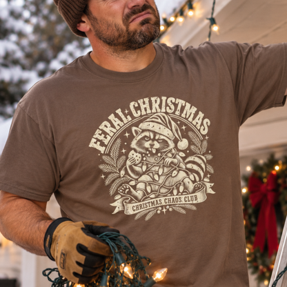 A man wearing a brown graphic t-shirt with a beige 'Feral Christmas' raccoon design, holding Christmas lights in a festive outdoor setting.