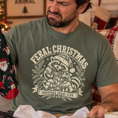 A man wearing an olive green graphic t-shirt with a cream-colored 'Feral Christmas' raccoon illustration, sitting in a cozy holiday home setting.