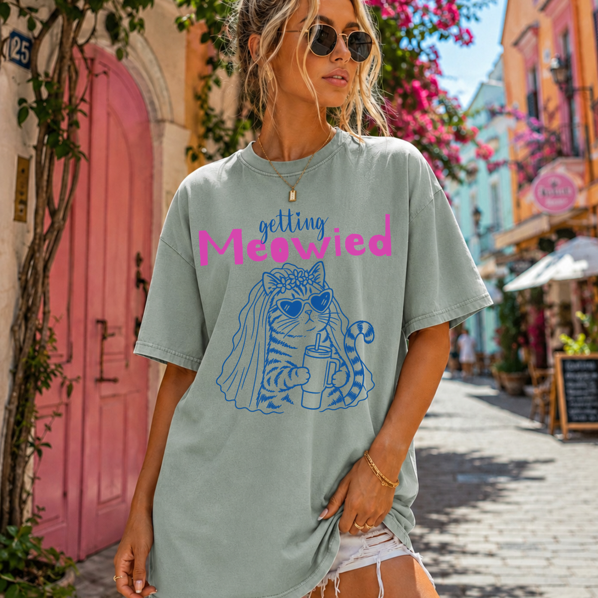 A woman wearing an oversized sage green graphic t-shirt with the text 'getting Meowed' and a blue cat illustration, standing on a sunny cobblestone street.