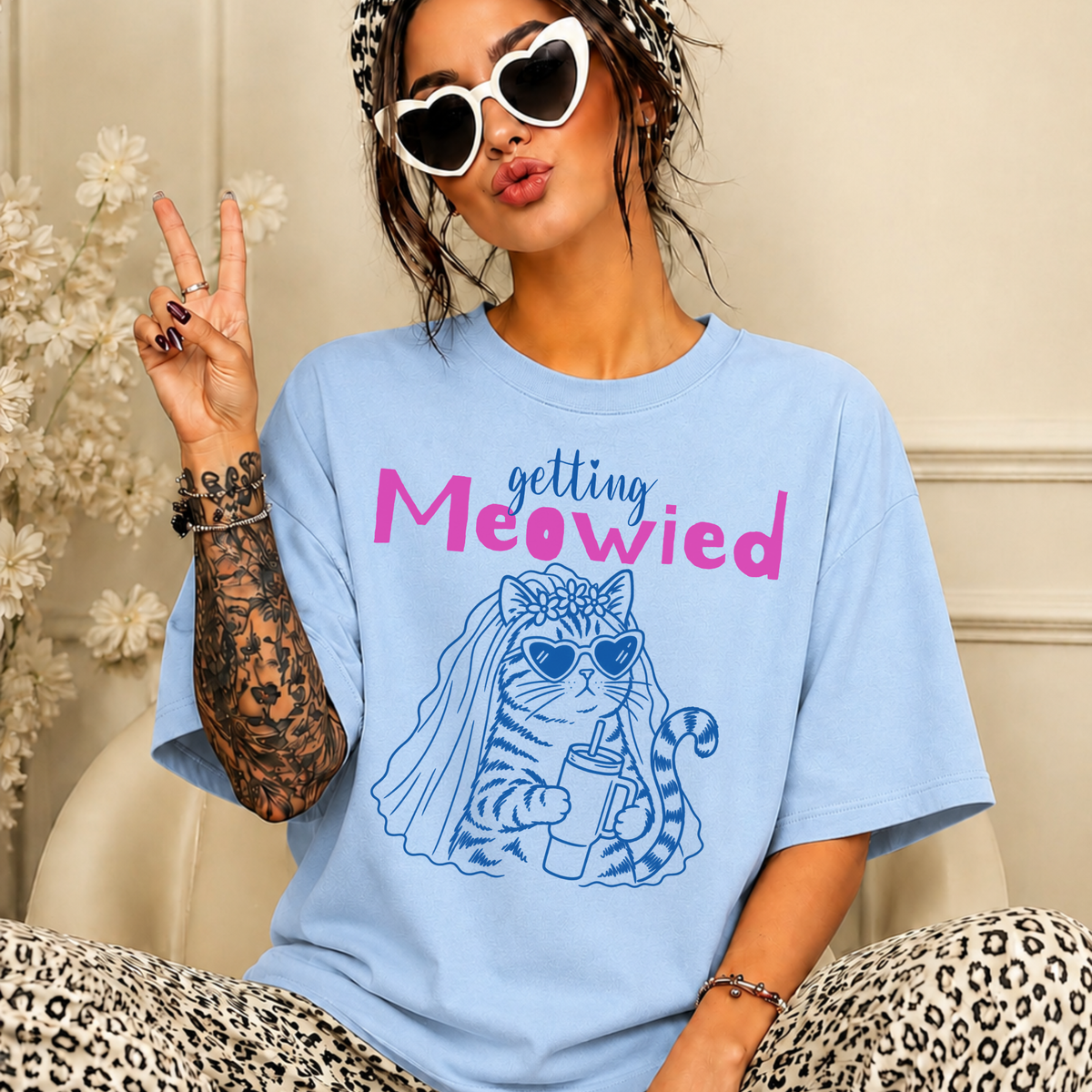 A woman wearing an oversized light blue graphic T-shirt with the text 'getting Meowied' and a blue cat illustration, posed indoors with leopard-print pants and white heart-shaped sunglasses.