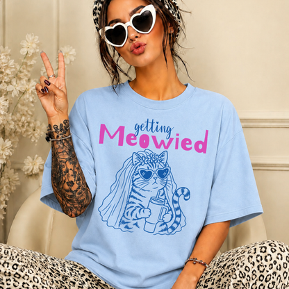 A woman wearing an oversized light blue graphic T-shirt with the text 'getting Meowied' and a blue cat illustration, posed indoors with leopard-print pants and white heart-shaped sunglasses.