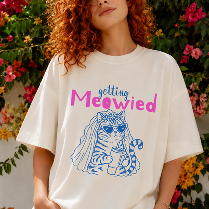 A woman wearing an oversized cream-colored graphic t-shirt with a blue and pink cat illustration and the text 'getting Meowied'. She is standing outdoors in front of a background of green foliage and pink flowers.