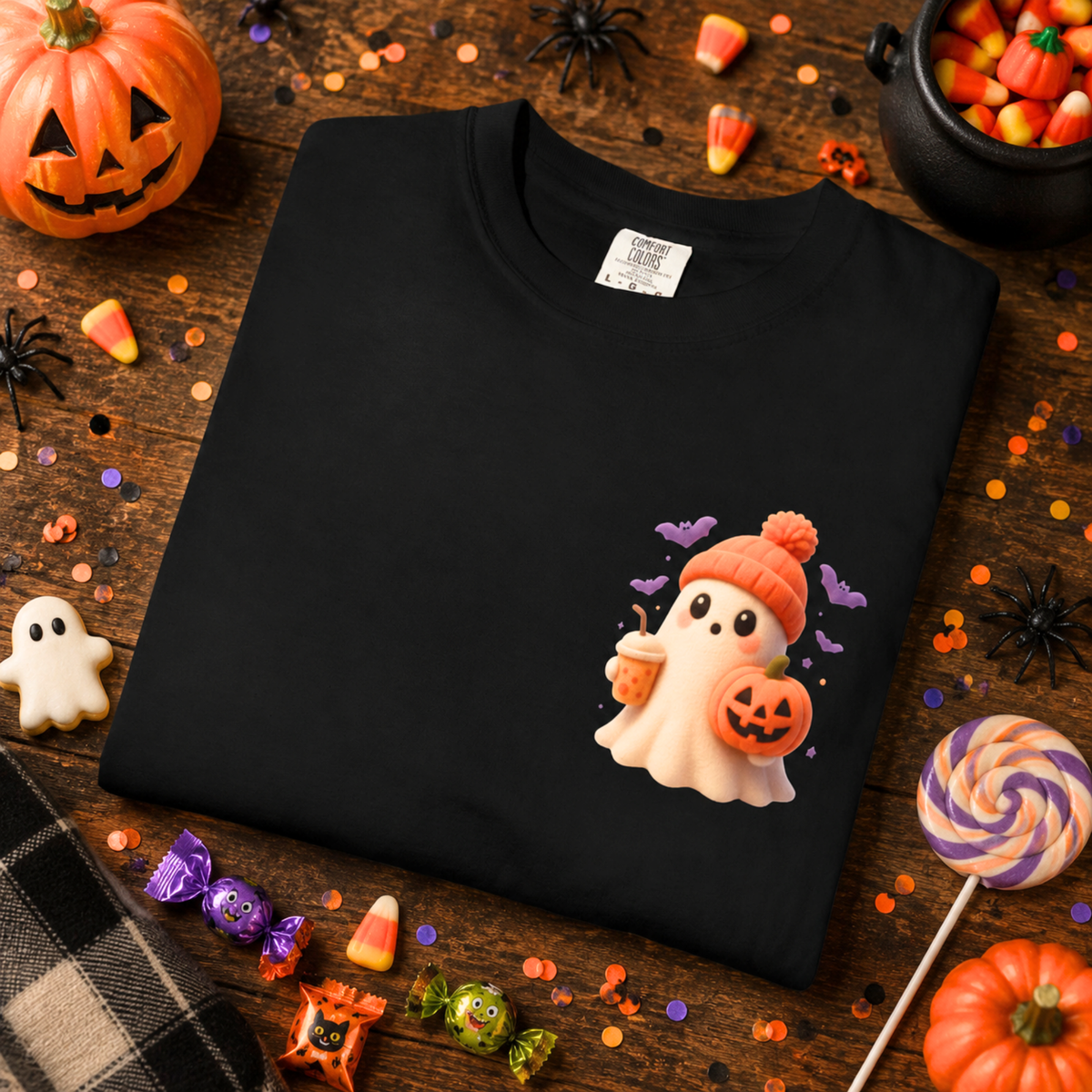 A folded black t-shirt with a cute cartoon ghost graphic wearing an orange beanie and holding a jack-o'-lantern, styled in a Halloween flat-lay with candy, pumpkins, and confetti on a wooden background.