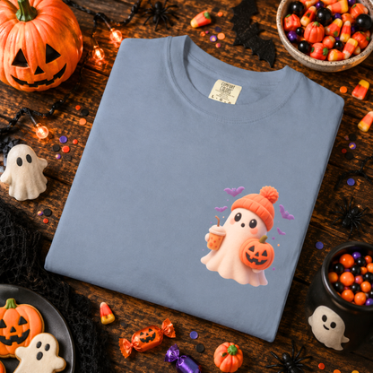 A folded light blue crewneck t-shirt with a cute cartoon ghost graphic, styled in a Halloween-themed flat lay on a dark wooden table surrounded by candy, pumpkins, and spider decorations.