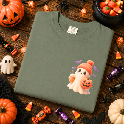 A folded sage green t-shirt with a cute cartoon ghost graphic wearing an orange hat and holding a jack-o'-lantern, displayed on a rustic wooden table decorated with Halloween props like pumpkins, candy corn, a cauldron, and black spiders.