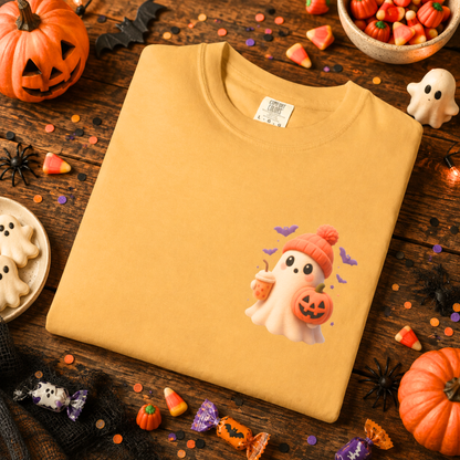 A folded mustard yellow crewneck sweatshirt with a cute cartoon ghost graphic on the chest, styled in a Halloween-themed flat lay with pumpkins, candy corn, and spider decorations.
