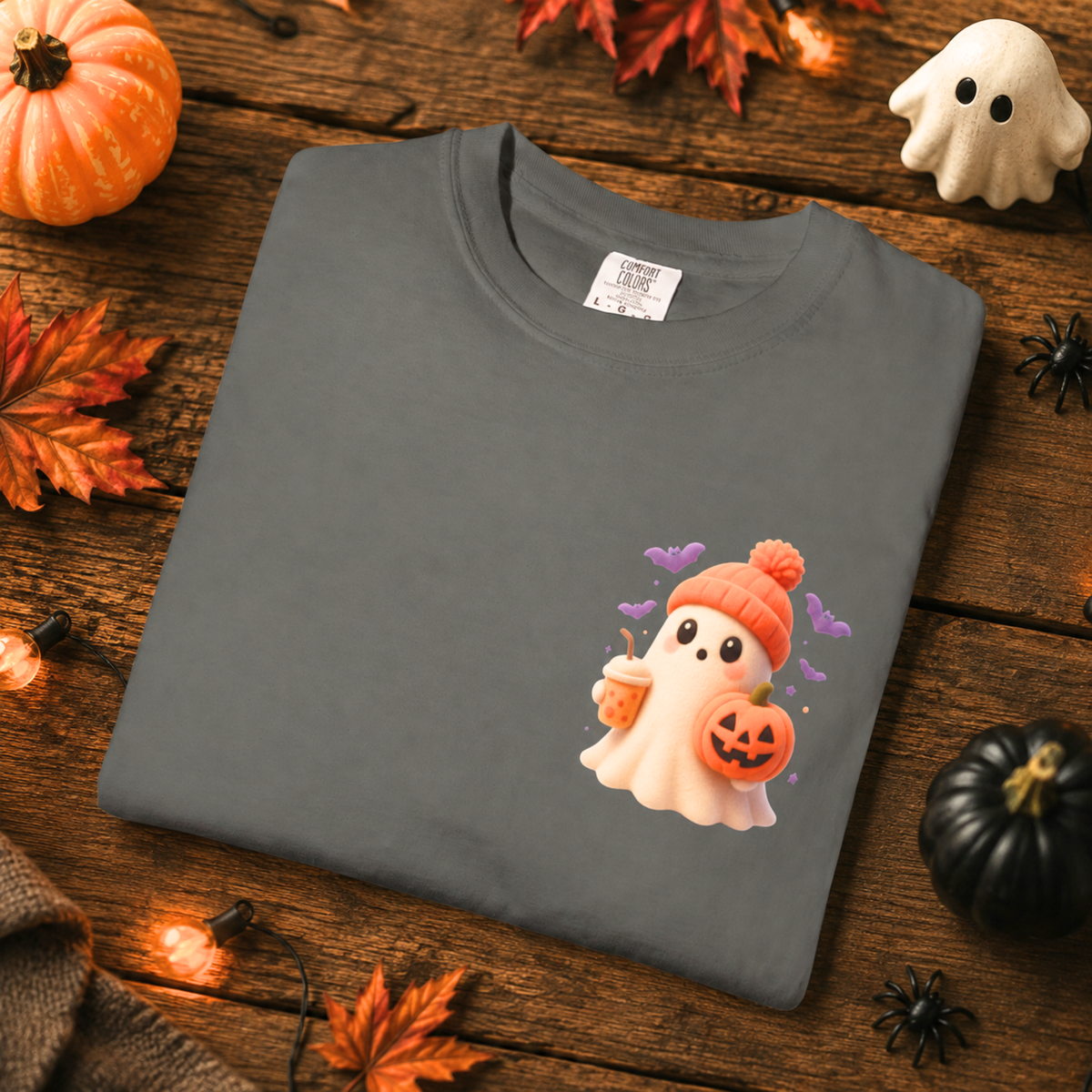 A folded dark gray t-shirt with a cute cartoon ghost wearing an orange beanie and holding a jack-o'-lantern, displayed on a rustic wooden table decorated with autumn leaves, pumpkins, and a small white ghost figurine.