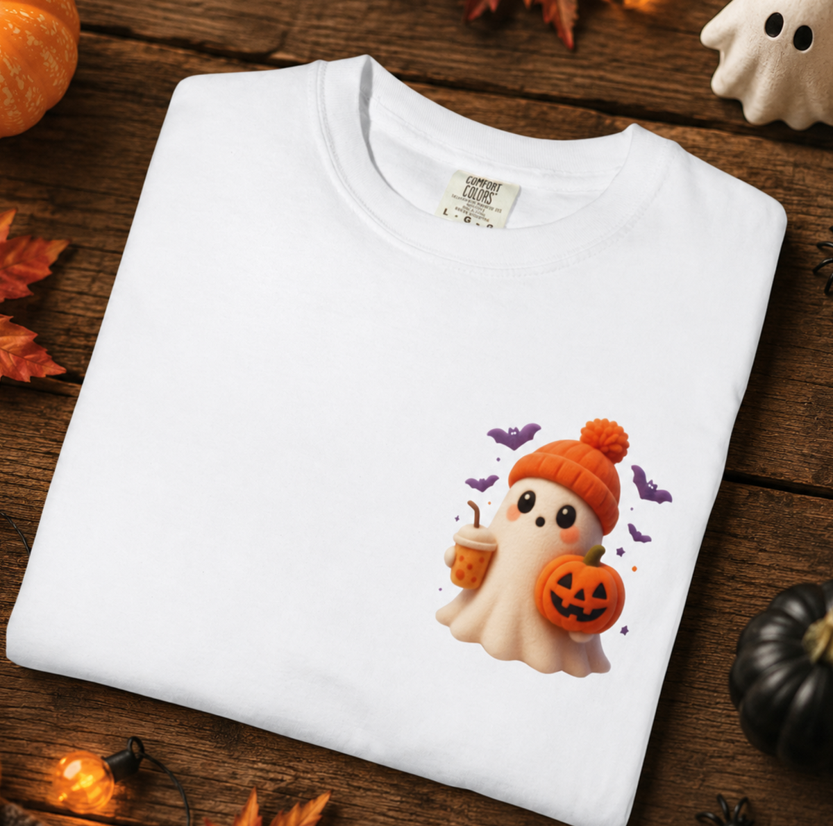 A white short-sleeve T-shirt laid flat on a rustic wooden surface, decorated with Halloween props including pumpkins, autumn leaves, ghost figurines, and string lights. The shirt features a cute cartoon ghost graphic wearing an orange beanie and holding a pumpkin cup.