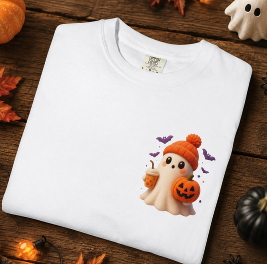 A white short-sleeve T-shirt laid flat on a rustic wooden surface, decorated with Halloween props including pumpkins, autumn leaves, ghost figurines, and string lights. The shirt features a cute cartoon ghost graphic wearing an orange beanie and holding a pumpkin cup.