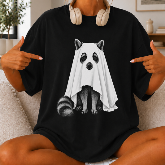 A person sitting on a couch wearing an oversized black T-shirt with a graphic of a raccoon covered in a white ghost. The person is pointing at the shirt with both hands, and beige headphones are hanging around the neck.