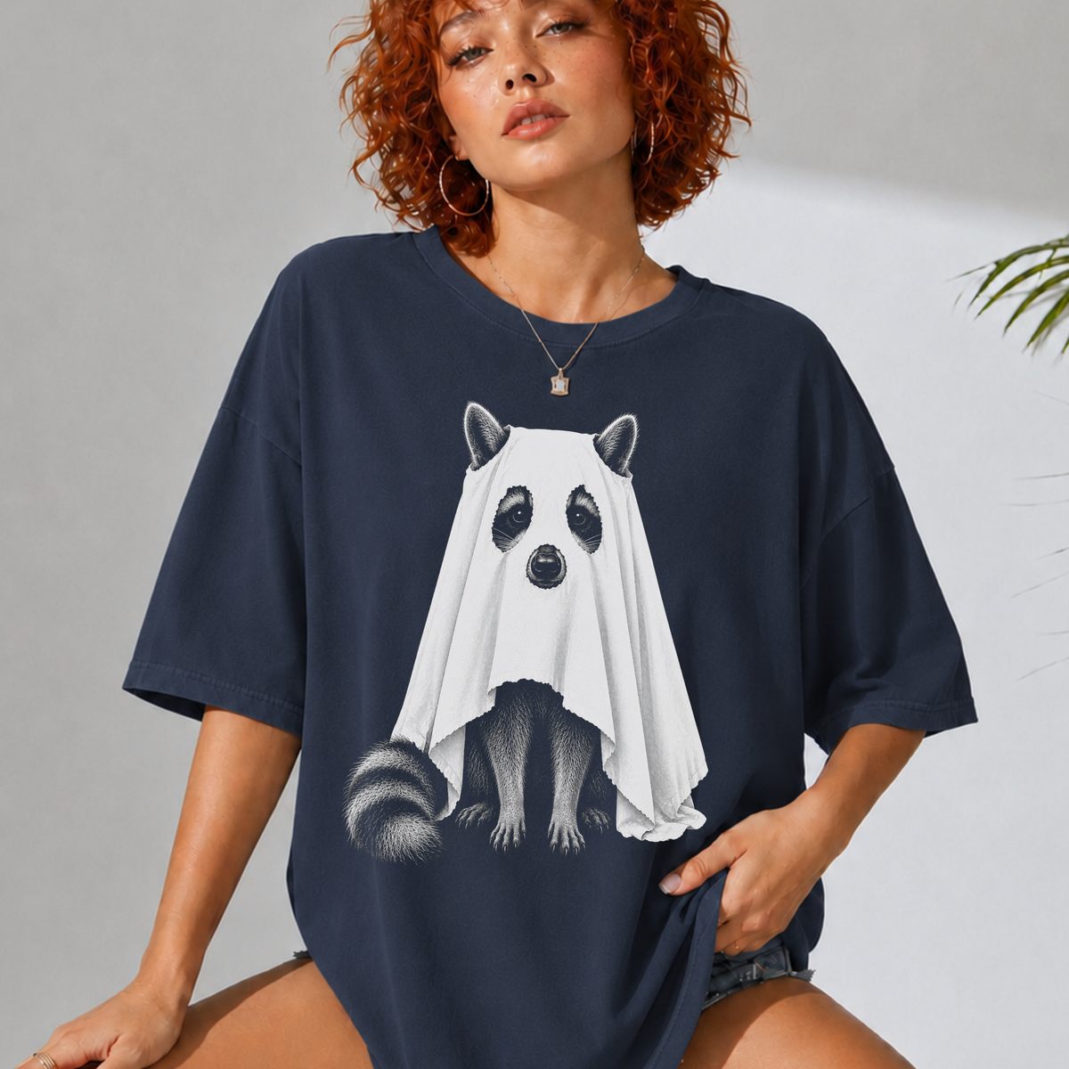 A woman wearing an oversized navy blue t-shirt with a graphic of a raccoon dressed in a white ghost, posed against a light gray wall.