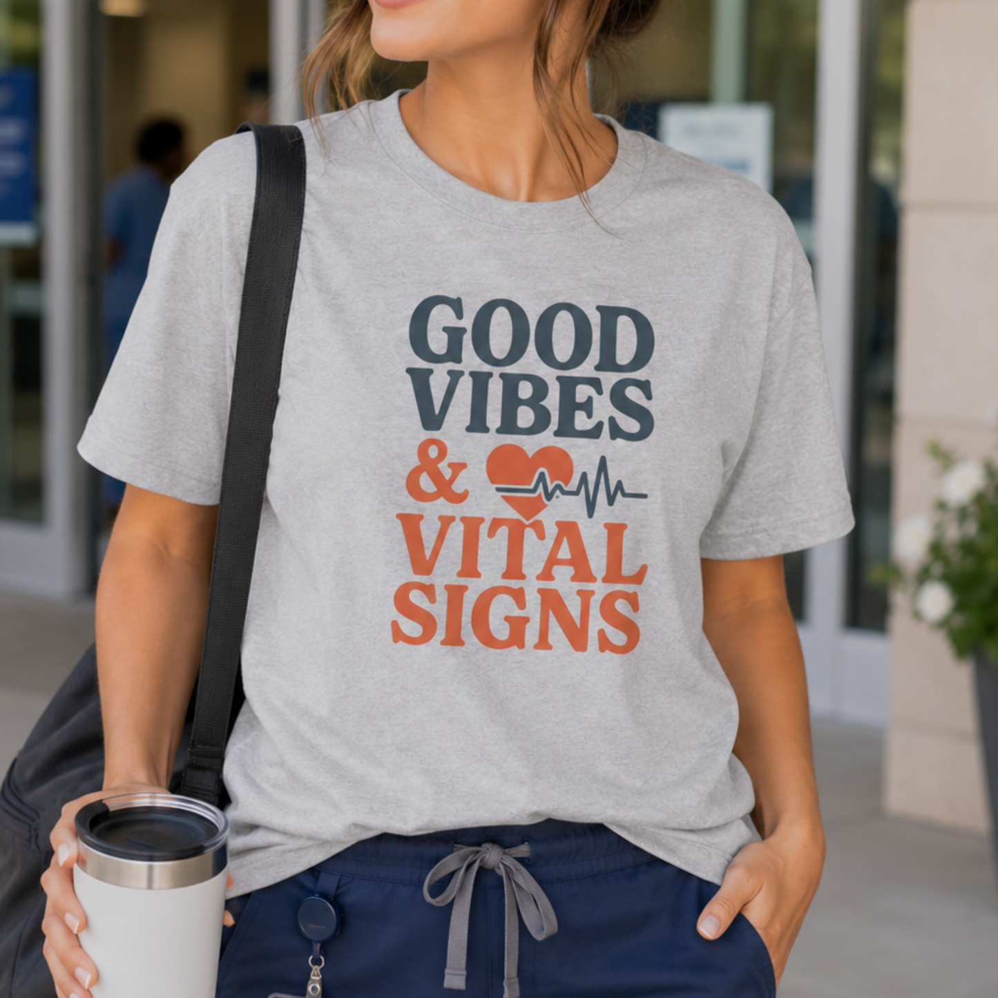 A woman wearing a heather gray graphic t-shirt with the text 'GOOD VIBES & VITAL SIGNS' in dark gray and orange, paired with navy blue sweatpants and carrying a black shoulder bag.