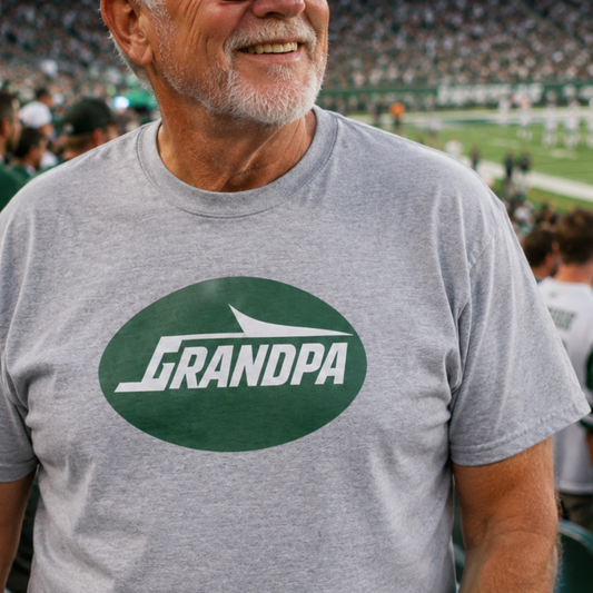 A smiling man at a sports stadium wearing a heather gray t-shirt with a green oval graphic that reads 'GRANDPA' in white.