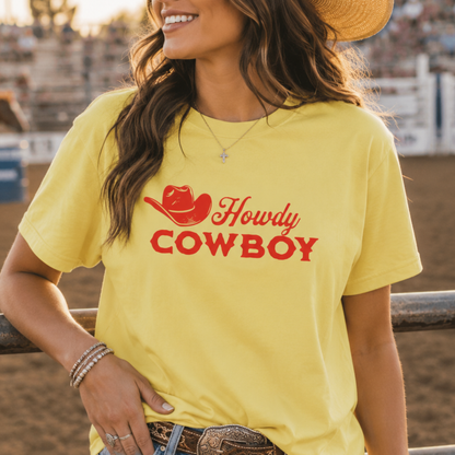 A woman wearing a bright yellow t-shirt with a red graphic that reads 'Howdy COWBOY' with a cowboy hat illustration, standing in an outdoor rodeo arena.