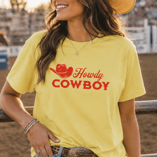 A woman wearing a bright yellow t-shirt with a red graphic that reads 'Howdy COWBOY' with a cowboy hat illustration, standing in an outdoor rodeo arena.