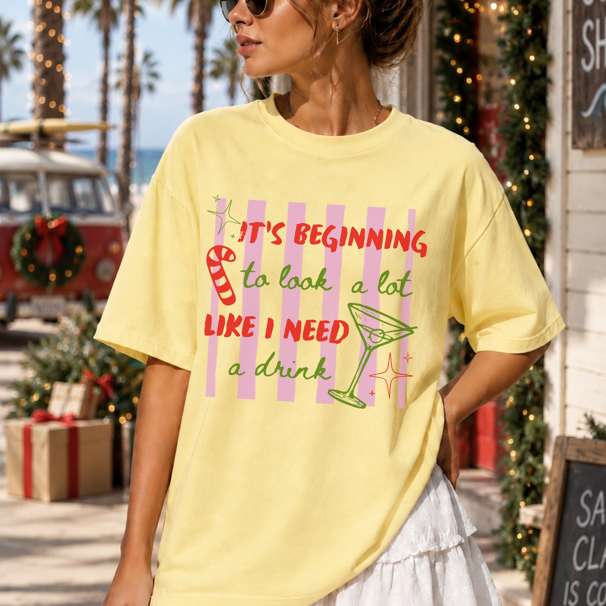 A woman wearing a pale yellow graphic t-shirt with a pink, red, and green holiday-themed print that reads 'IT'S BEGINNING to look a lot LIKE I NEED a drink'. The background is a festive outdoor street scene with palm trees, a vintage van, and holiday decorations.