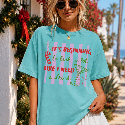A woman wearing an oversized turquoise graphic t-shirt with a holiday-themed drink design and text, photographed outdoors on a sunny beachside walkway with palm trees and holiday decorations in the background.