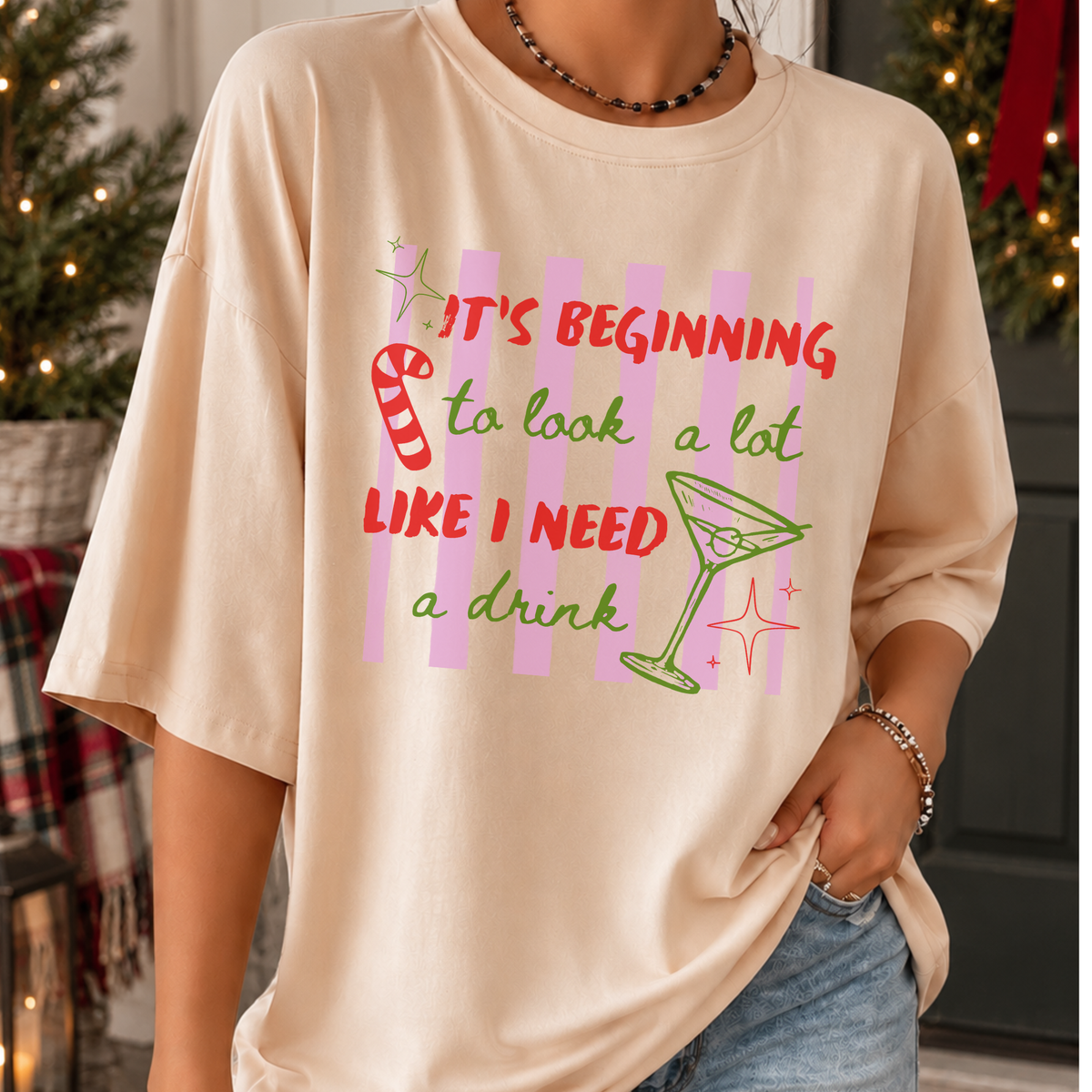 A person wearing a light beige oversized graphic T-shirt with a Christmas-themed slogan in red and green text, pink stripes, a candy cane, and a martini glass. The background shows a festive holiday setting with a decorated Christmas tree and warm lights.