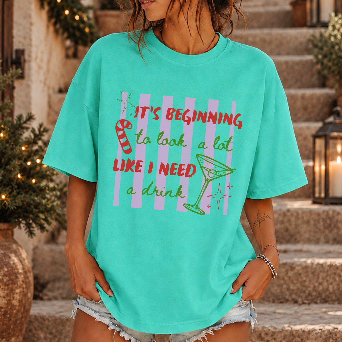 A woman wearing a turquoise graphic t-shirt with a holiday-themed print that reads 'IT'S BEGINNING to look a lot LIKE I NEED a drink' alongside a martini glass and candy cane illustration, set against a background of stone steps and holiday decorations.