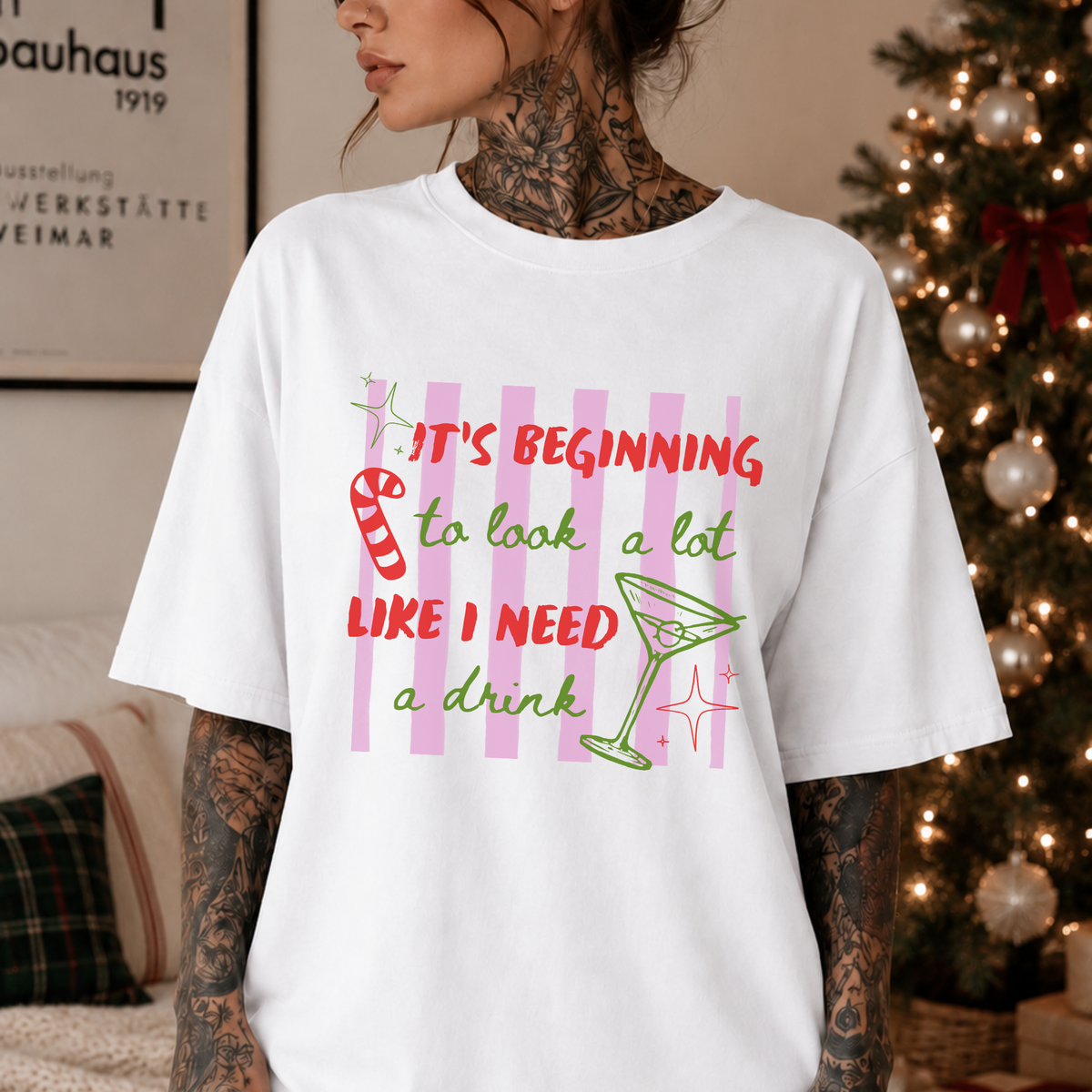 A woman wearing a white graphic t-shirt with a colorful text design that reads 'IT'S BEGINNING to look a lot LIKE I NEED a drink' with a candy cane and martini glass illustration, set against a cozy indoor background with a lit Christmas tree.