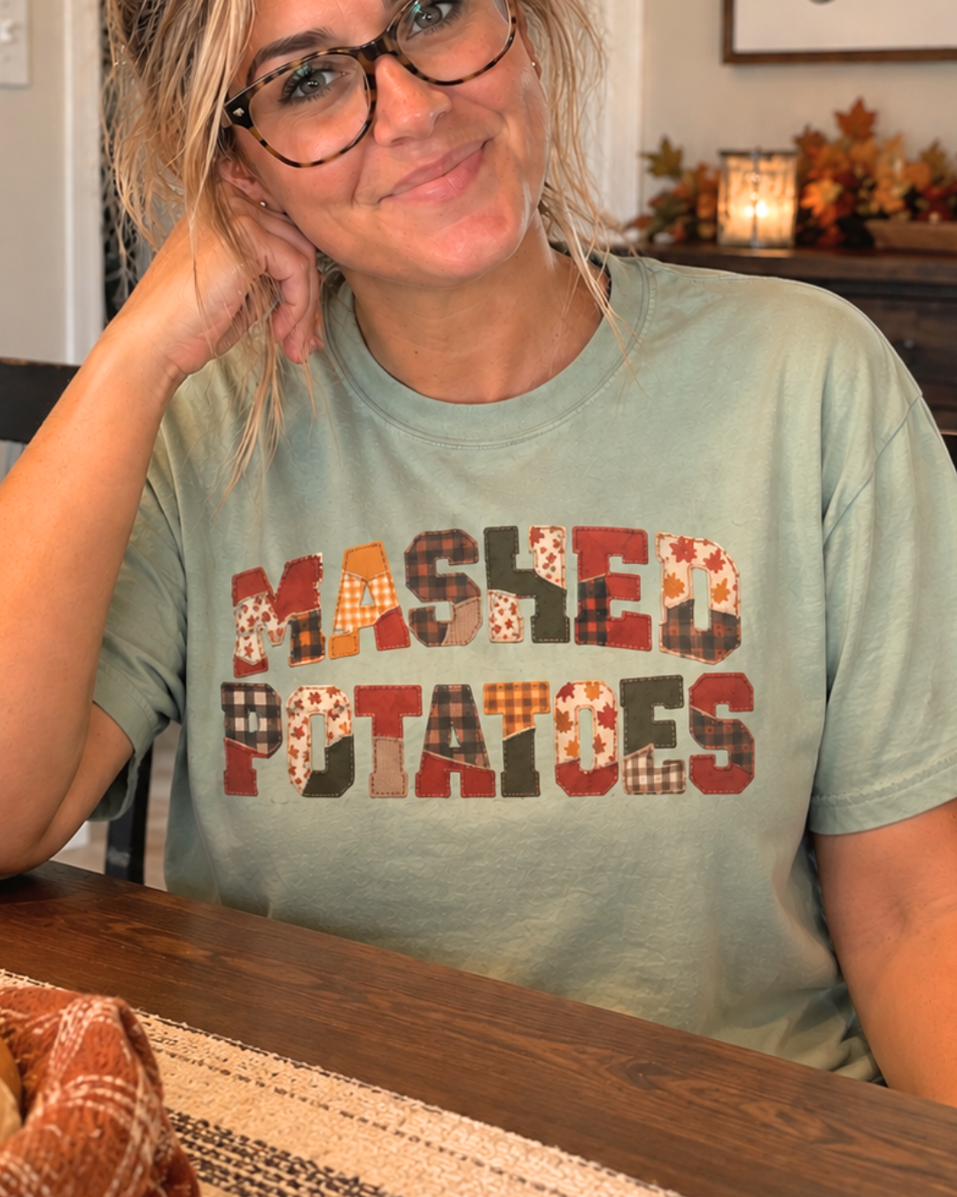 A woman with glasses smiling while wearing a sage green t-shirt with 'MASHED POTATOES' printed in a patchwork pattern of autumn colors, sitting at a wooden table.