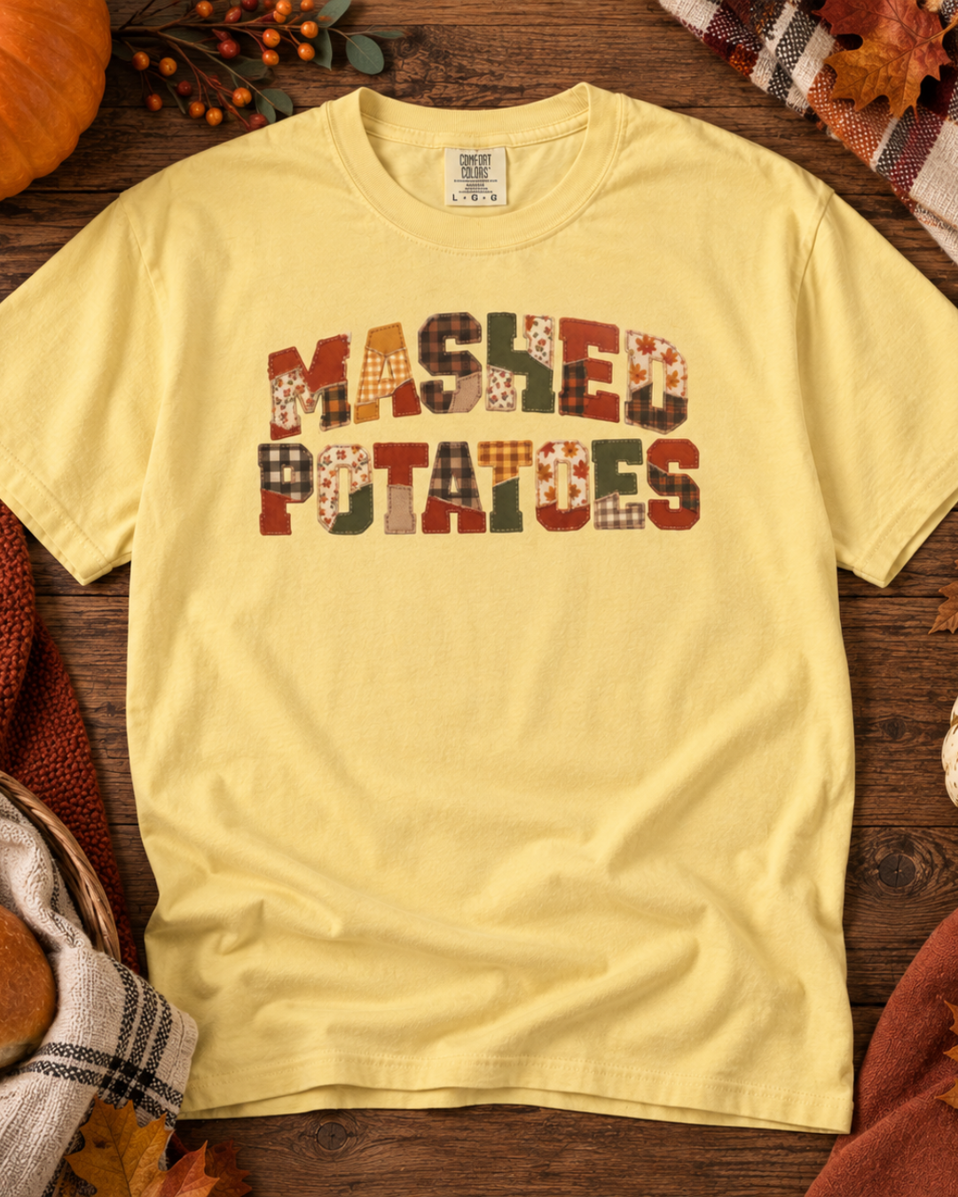 A pale yellow short-sleeve t-shirt laid flat on a rustic wooden surface, featuring the phrase 'MAShED POTATOES' in a patchwork-style multicolor graphic across the chest. The shirt is styled with autumn props including a pumpkin, plaid fabric, and leaves.