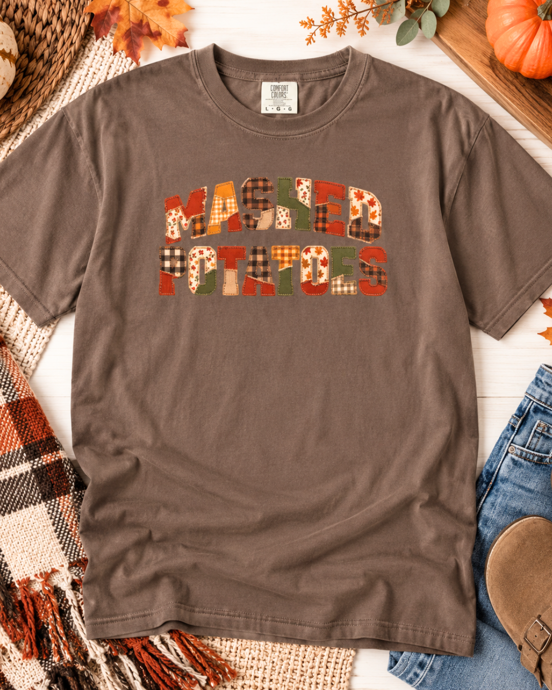 A brown short-sleeve t-shirt laid flat on a white wood surface, featuring the phrase 'MASHED POTATOES' in a patchwork plaid pattern of orange, green, red, and brown tones. The shirt is styled with autumn props including a plaid blanket, a pumpkin, leaves, and denim jeans.