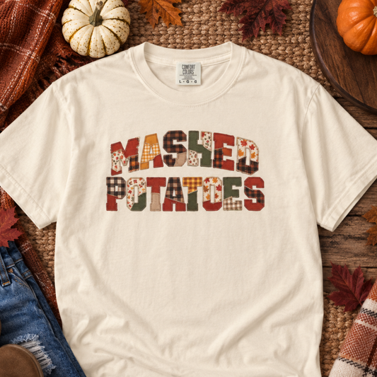 An off-white short-sleeve t-shirt with a graphic reading 'MASHED POTATOES' in a patchwork pattern of autumn colors, styled in a flat lay with jeans, a plaid scarf, pumpkins, and fall leaves.