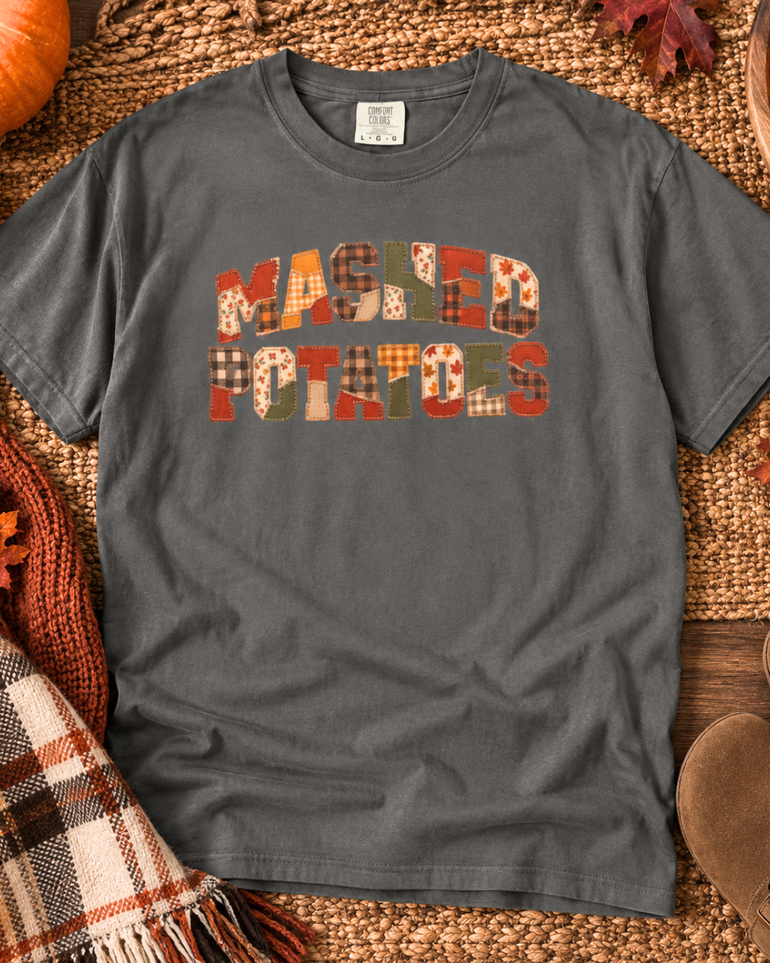 A dark gray crewneck t-shirt laid flat on a woven mat, featuring the text 'MASKED POTATOES' in a patchwork plaid pattern of orange, red, green, and brown tones. The shirt is styled with autumn props including a pumpkin, a knit scarf, and fall leaves.