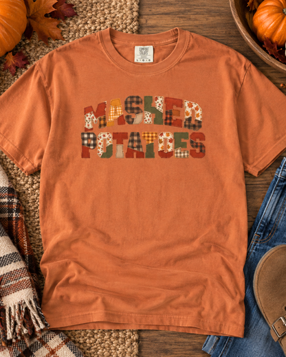 An orange crewneck t-shirt featuring a 'Mashed Potatoes' graphic made of various autumn-themed patchwork patterns, styled in a flat-lay with pumpkins, a plaid scarf, and blue jeans.