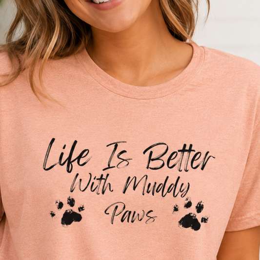 A close-up of a woman wearing a heathered peach-pink t-shirt with the black printed text 'Life Is Better With Muddy Paws' and paw prints.