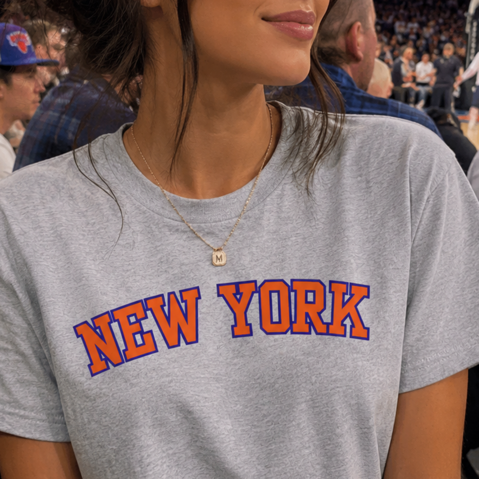 A close-up of a person wearing a heather gray t-shirt with 'NEW YORK' printed in orange and blue varsity lettering across the chest, set against a crowded indoor background.