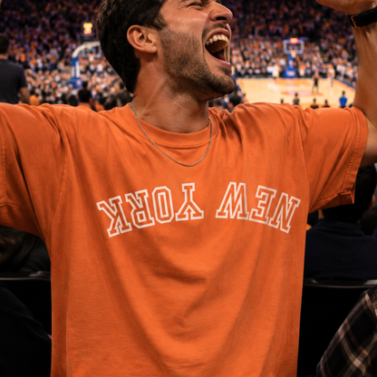 A man cheering at a basketball game wearing an orange t-shirt with 'NEW YORK' printed in white arched text across the chest.