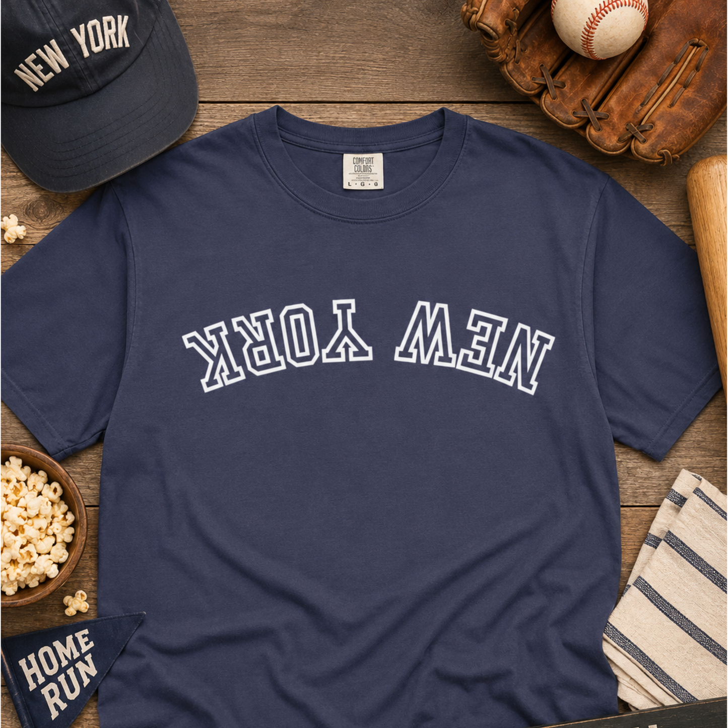 A navy blue crewneck t-shirt with 'NEW YORK' printed in white varsity block letters across the chest, styled in a flat lay with baseball-themed props including a cap, baseball, glove, popcorn, and a striped towel on a wooden surface.