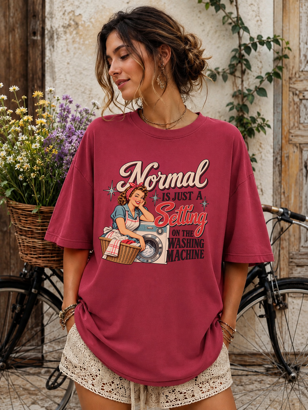 A woman wearing a relaxed-fit burgundy graphic t-shirt with a retro washing machine illustration and the text 'Normal is just a selling point on the washing machine'. She is paired with cream lace shorts, standing in front of a rustic wooden door and a bicycle.