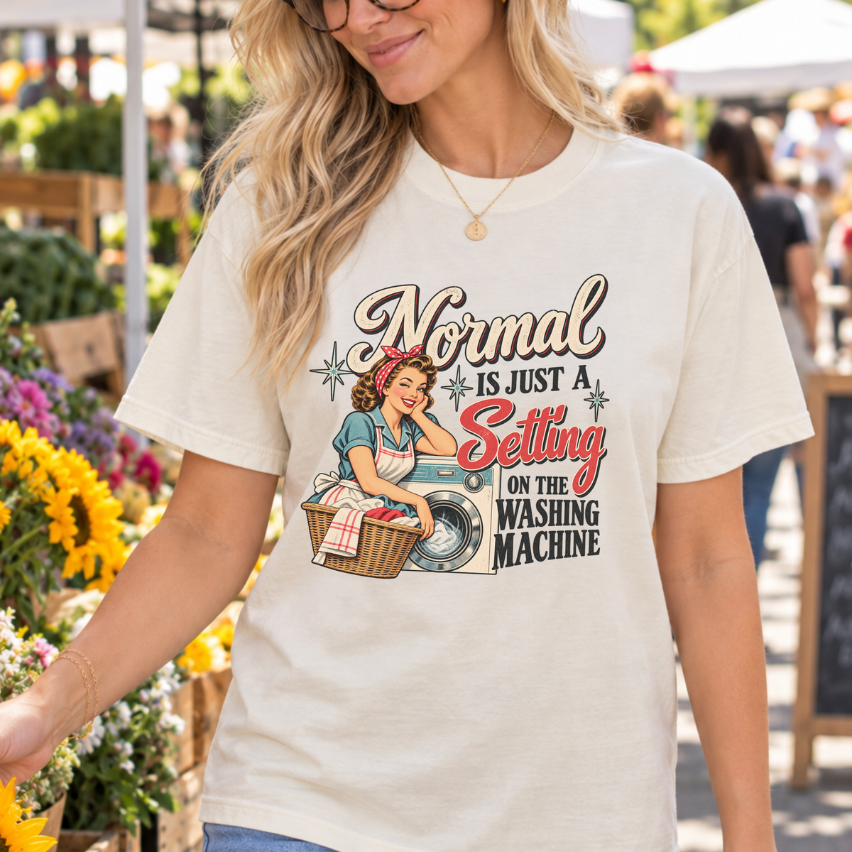 A woman wearing a cream-colored graphic t-shirt with a retro-style illustration and the text 'Normal is just a Setting on the Washing Machine' at an outdoor flower market.