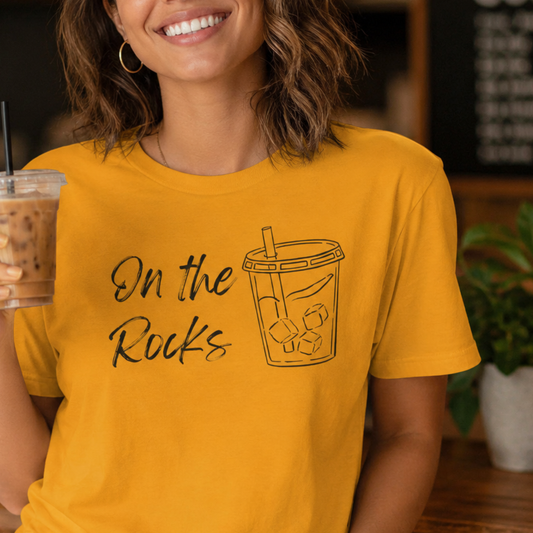 A woman wearing a mustard yellow graphic t-shirt with the text 'On the Rocks' and a black outline drawing of a iced coffee cup. She is holding an iced coffee in a cafe setting.