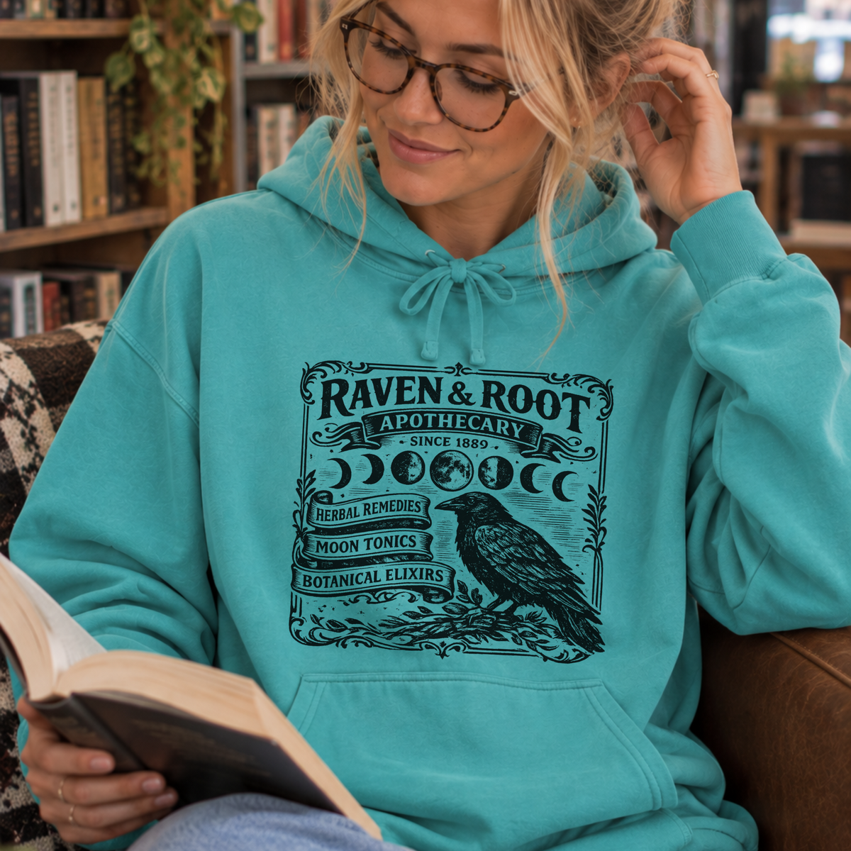 A woman wearing a turquoise hoodie with a black 'Raven & Root Apothecary' graphic sits in a cozy indoor setting, reading a book.