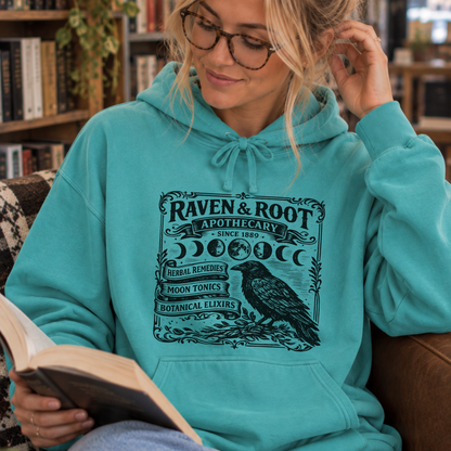 A woman wearing a turquoise hoodie with a black 'Raven & Root Apothecary' graphic sits in a cozy indoor setting, reading a book.